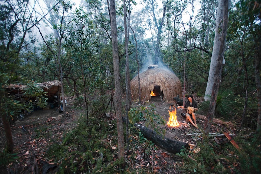 An woman sits by a fire in front of a grass hut, surrounded by forest.