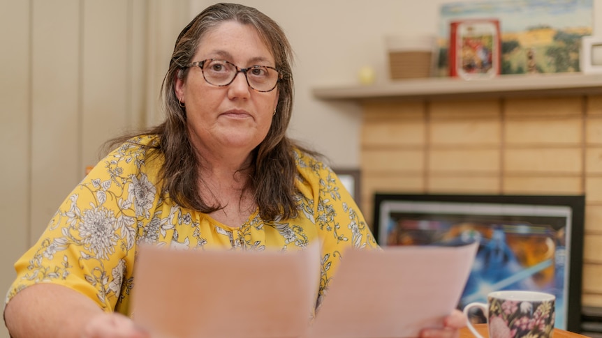 Bespectacled woman with brown hair and yellow top sitting at kitchen table holding power bills
