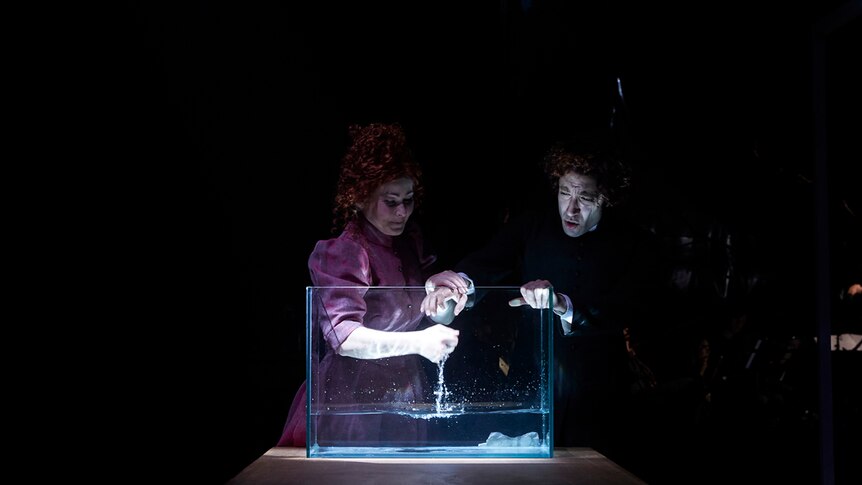 Man and woman dressed in colonial era costumes reaching into fishtank with some water on a dimly lit set.
