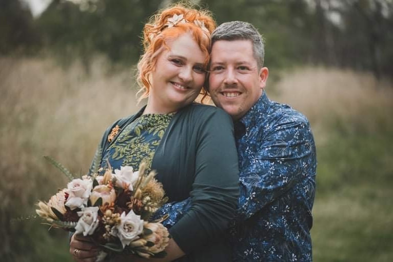 Un homme en chemise bleue se tient avec ses bras autour d'une femme en robe verte, tenant un bouquet de fleurs.