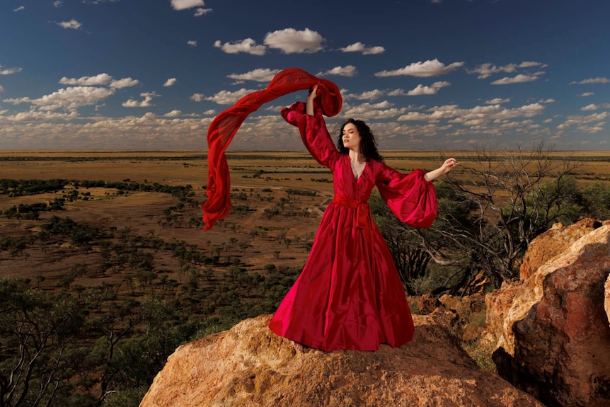 A woman in a red gown stands on the edge of a rock cliff with her arms outstretched.