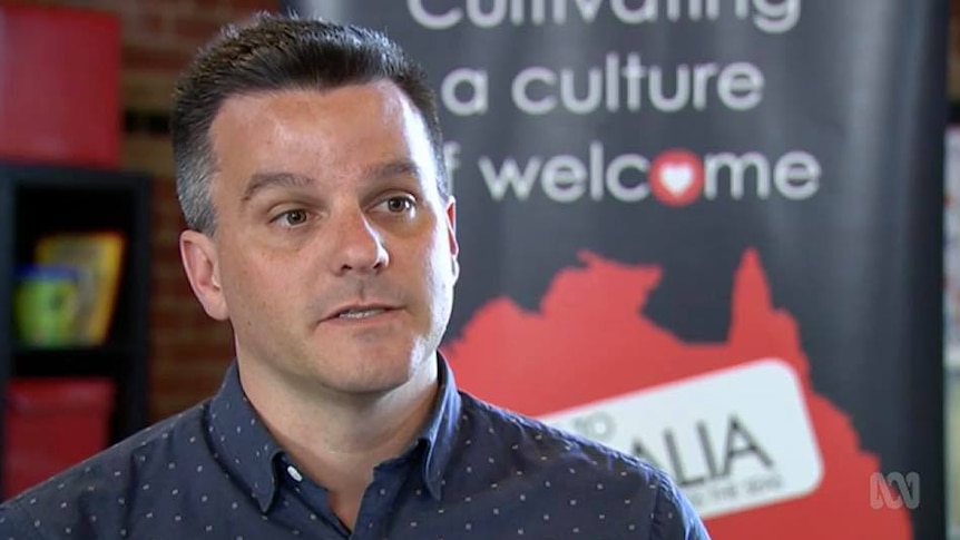 A man with short black and grey hair sits in front of a Welcome to Australia banner.