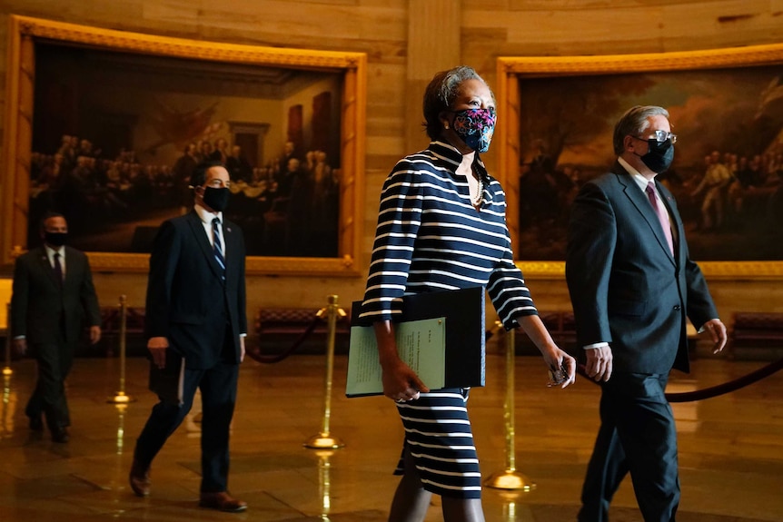 A black woman wearing a mask walks next to a white man also in a mask through a decadent hall. Two suited men follow.