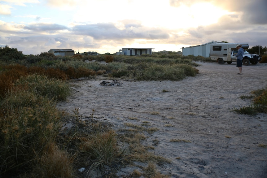 A sandy camp site at dusk, with tin shacks and a man and his vehicle in the background.