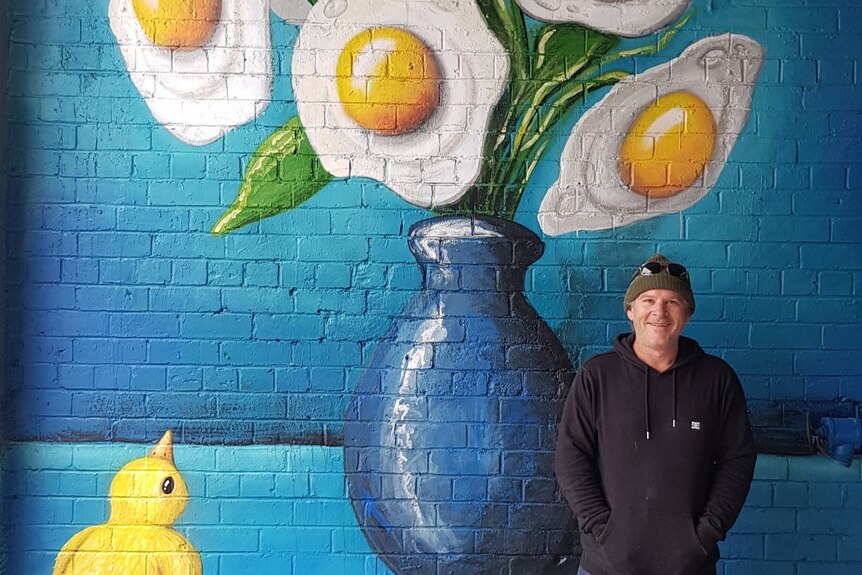 Artist Tank, wearing a black hoodie and dark beanie, stands in front of a wall mural.
