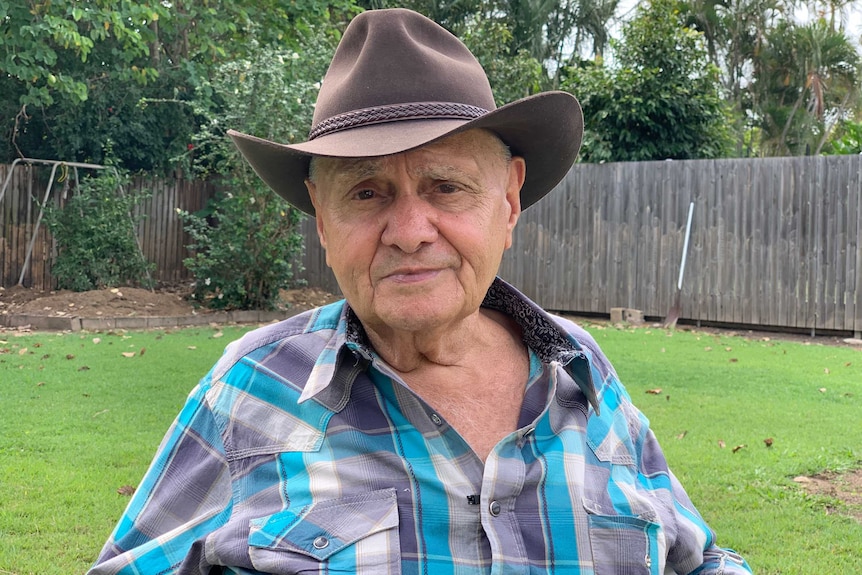 An elderly man in a checked shirt and broad hat sits in his backyard with grass and a fence in the background