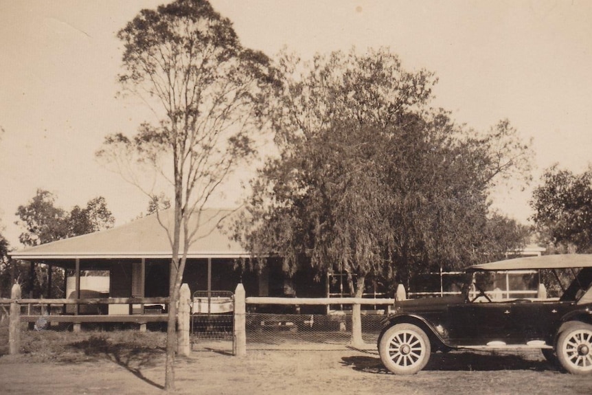 Black and white image of an old car in front of a homestead surrounded by trees.