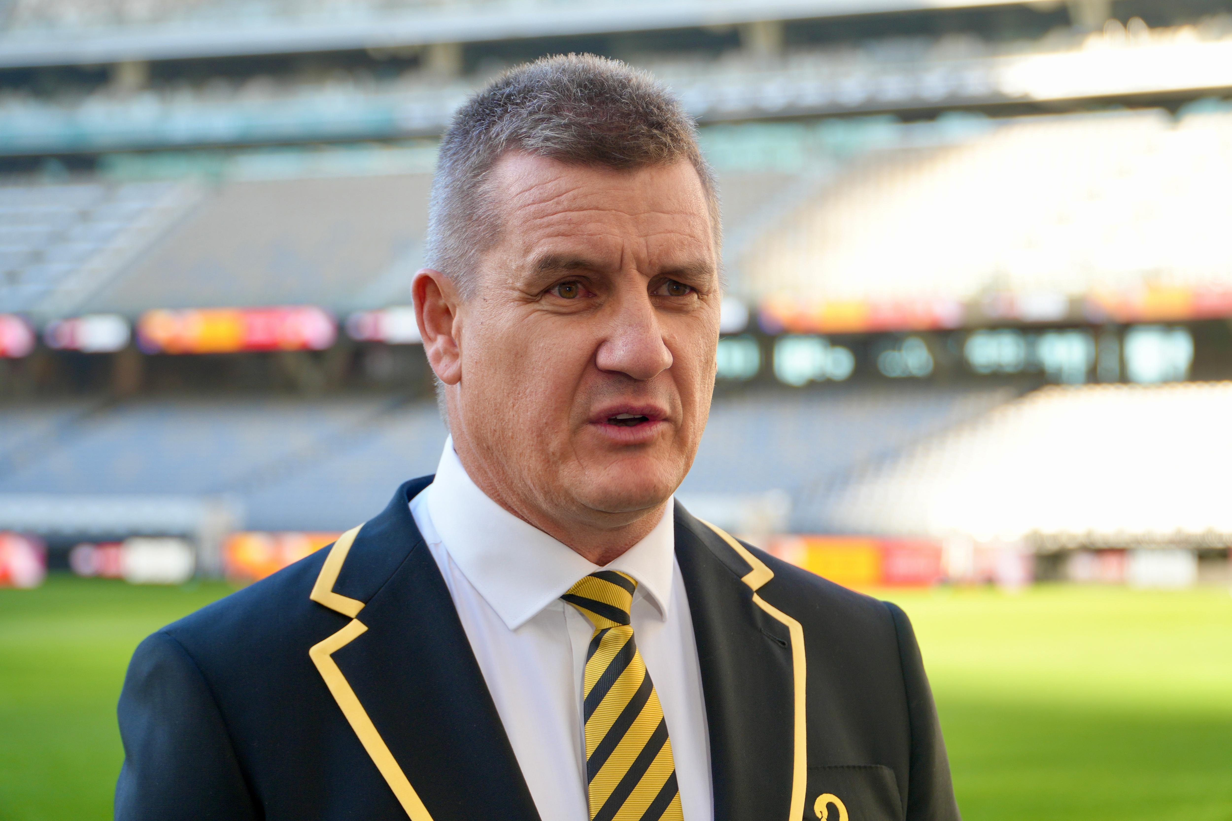 Glen Jakovich stands speaking while wearing a blazer and tie. 