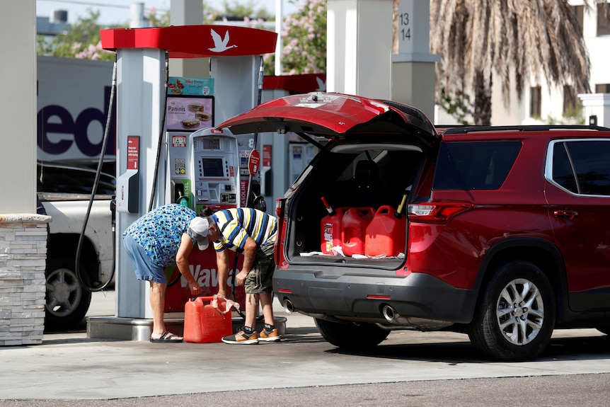 A couple fills up a red container with petrol. Three more containers are visible in their car boot.