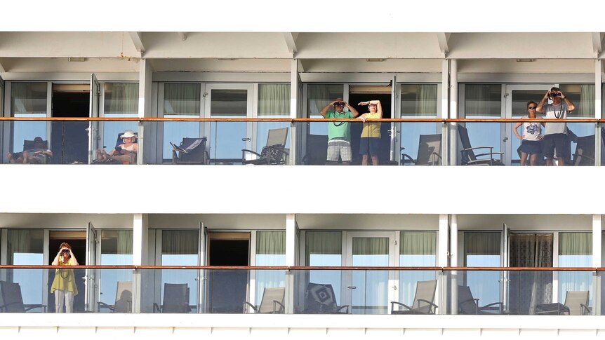 Passengers stand on the balconies of their rooms on a cruise ship. Some are using binoculars