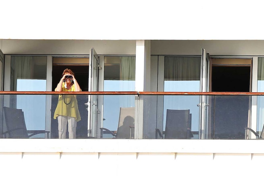 Passengers stand on the balconies of their rooms on a cruise ship. Some are using binoculars