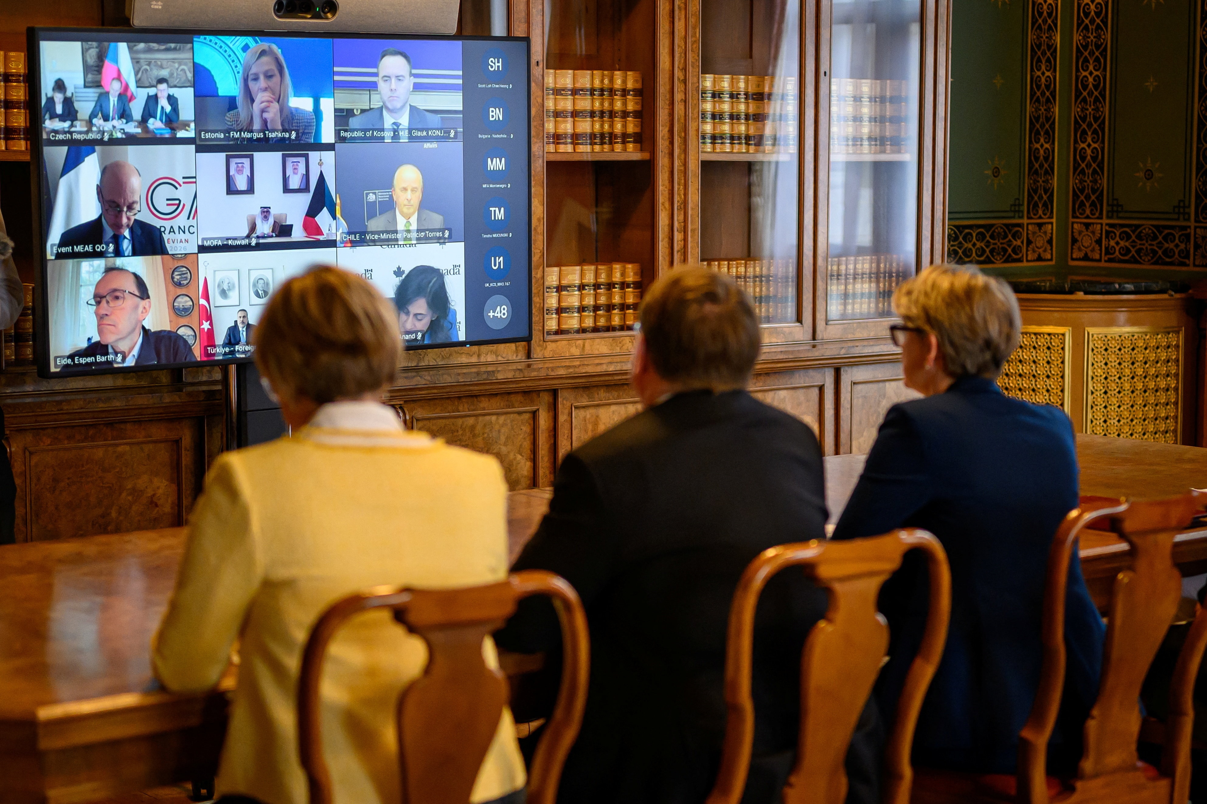 Τρεις άνθρωποι έχουν την πλάτη τους στην κάμερα μπροστά σε μια οθόνη με τους ηγέτες του κόσμου σε μια κλήση συνδιάσκεψης.