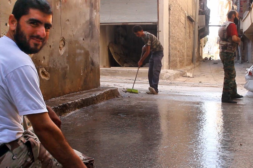 A man holds a hose in the street as a teenager sweeps up behind and another mans stands by with binoculars.