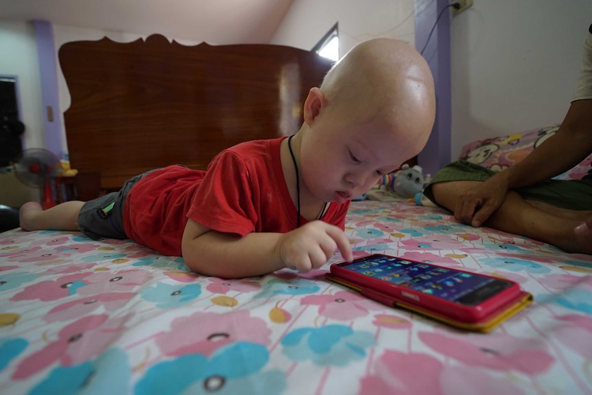 Gramy wears a red shirt and grey cargo shorts as hey lays on a bed and plays with an iphone in a red case.