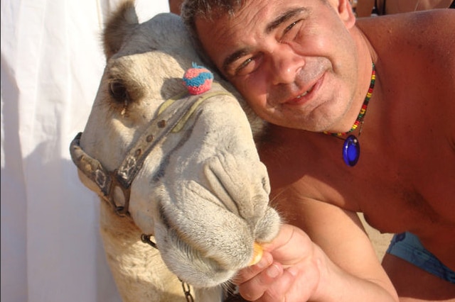 Oleg poses with a camel, either feeding it or holding its halter. He wears brightly-coloured beads around his neck and no shirt.