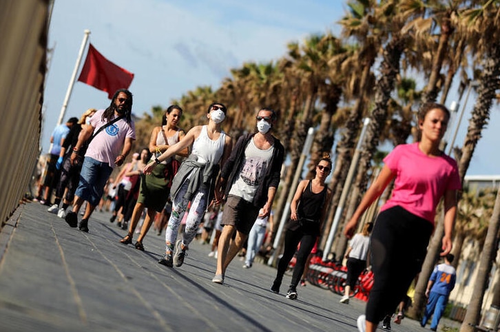 Tens of people walk along the pavement with palm trees overhead. Some wear masks, the sky above is blue.