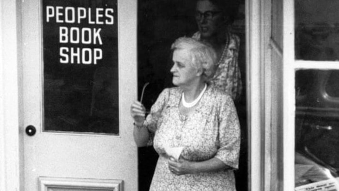 Black and white image of Anne Neill with neat hair and long dress, holding glasses, exiting door of the 'People's Book Shop'.