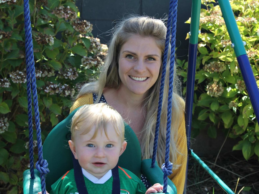 A woman in a yellow jumper crouches behind her smiling young son in a swing seat.