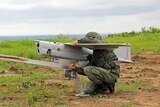 A person in military uniform crouched next to a drone