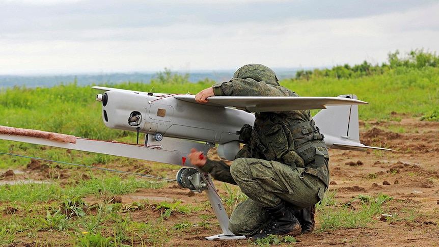 A person in military uniform crouched next to a drone