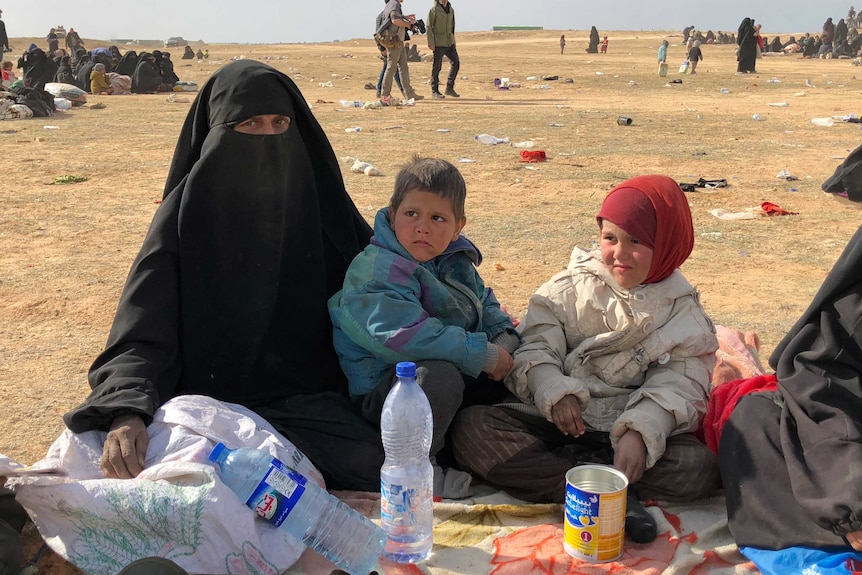A woman in Islamic clothing sits in an open area on a rug with two young children