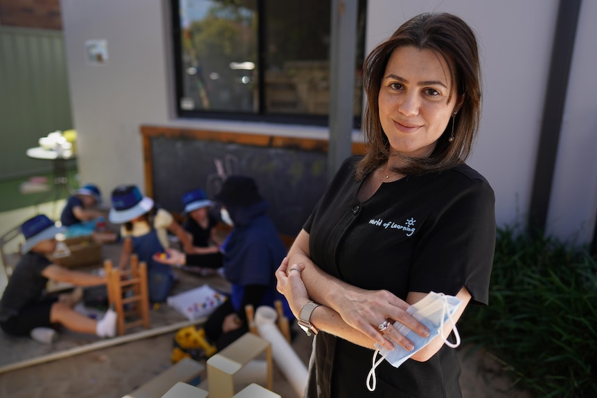 Graziela stands with her arms crossed in a childcare uniform, while children are seen playing the background.