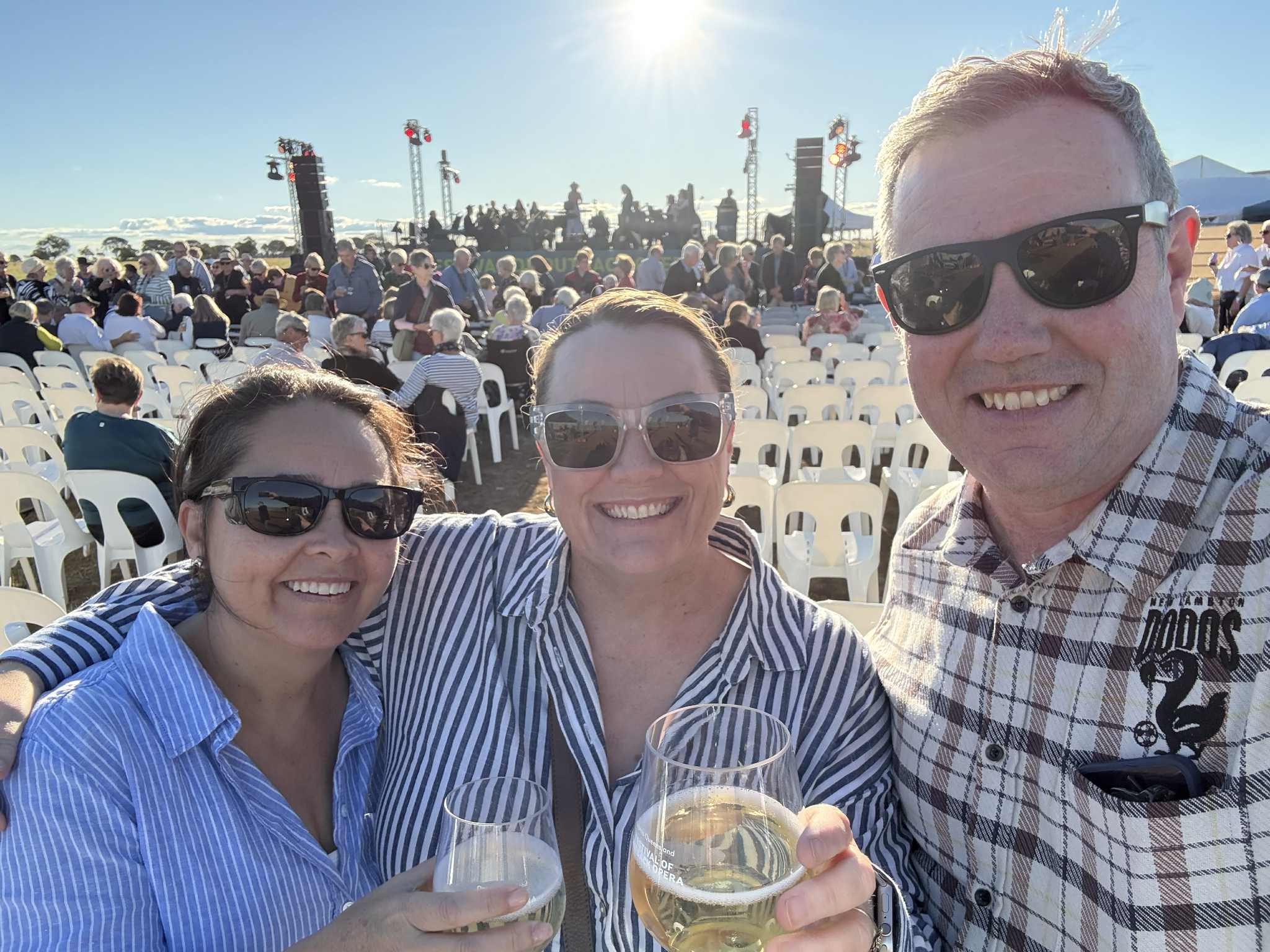 Samantha se para en medio de dos personas y se toma una selfie con un vino frente a un escenario y asientos. 