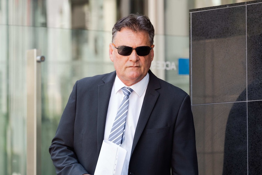 A man in a black suit and wearing black sunglasses walks out of a court building