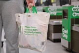 A person wearing grey trackpants holds a bulging reusable Woolworths-branded plastic bag.  