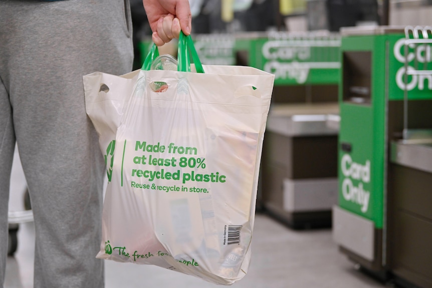 Osoba w szarych dresach trzyma puchową plastikową torbę wielokrotnego użytku z napisem Woolworths.  