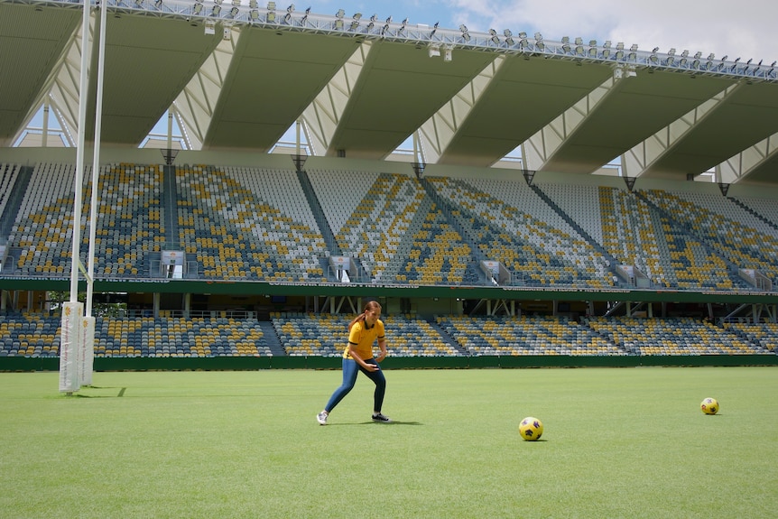A young girl wearing a green and gold jersey and jeans kicks a soccer-ball on the field by rugby league posts. 
