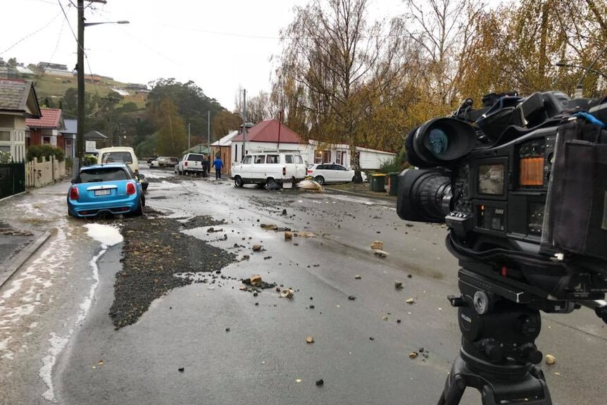 TV camera on tripod in street with water rushing down footpath and road broken up.