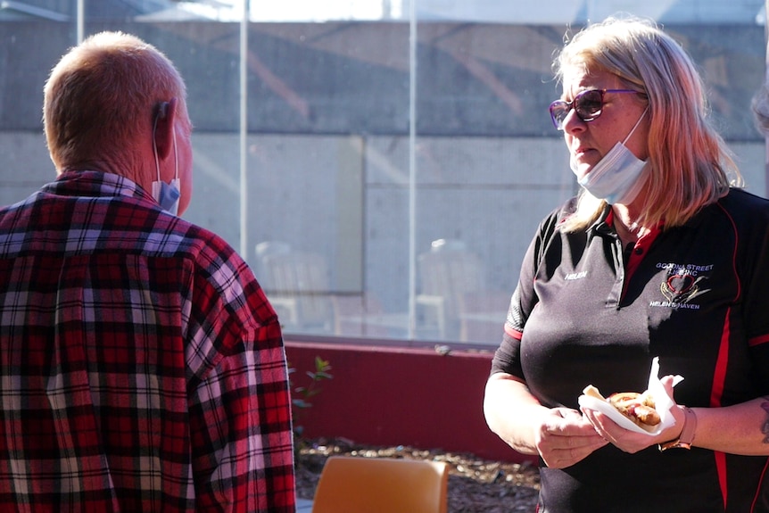 Woman stands holding a sausage sandwich while talking to a man wearing a mask.