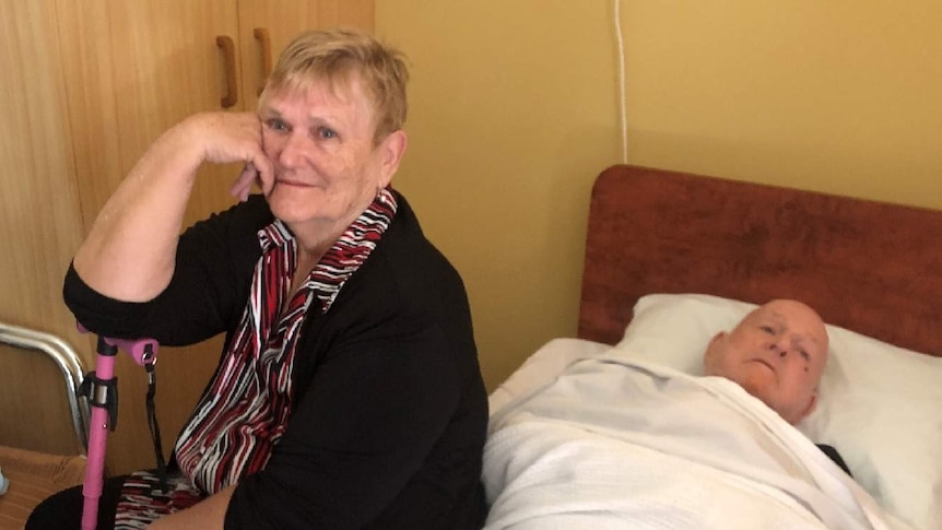 Lorraine Cook sits with her husband John who is laying on a bed.