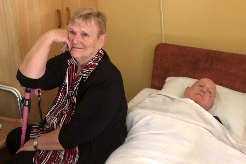 Lorraine Cook sits with her husband John who is laying on a bed.