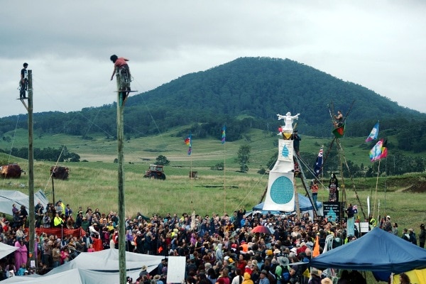 A crowd of people, some atop poles, in the countryside, with a green, forested hill in the background.