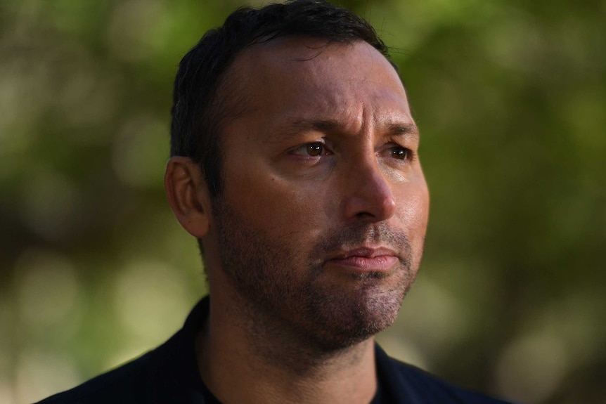 A man against a leafy background looks serious