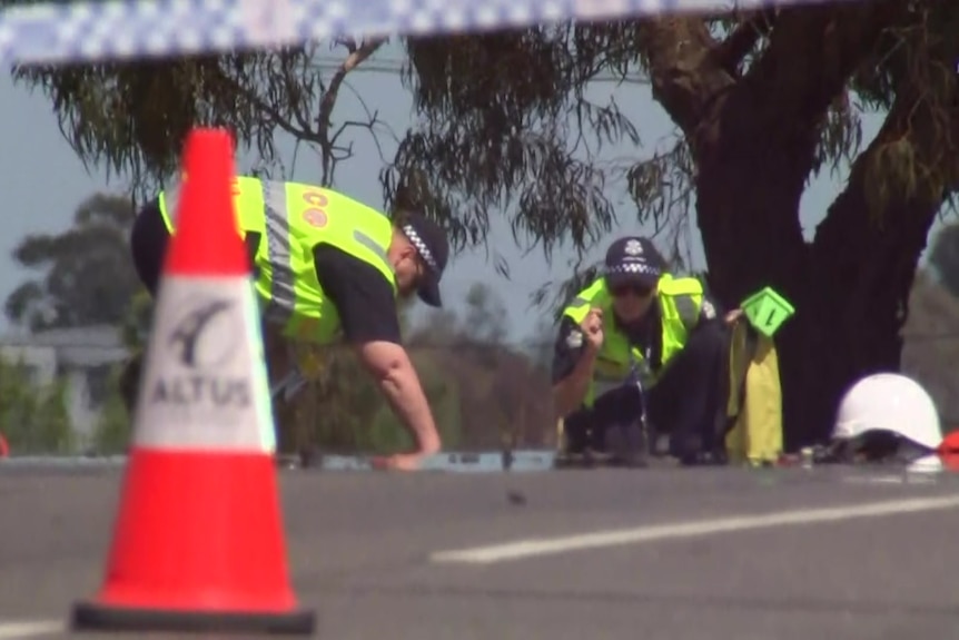 Two police officers in hi-vis vests crouch down as they survey evidence on the road around the crash site, behind police tape.