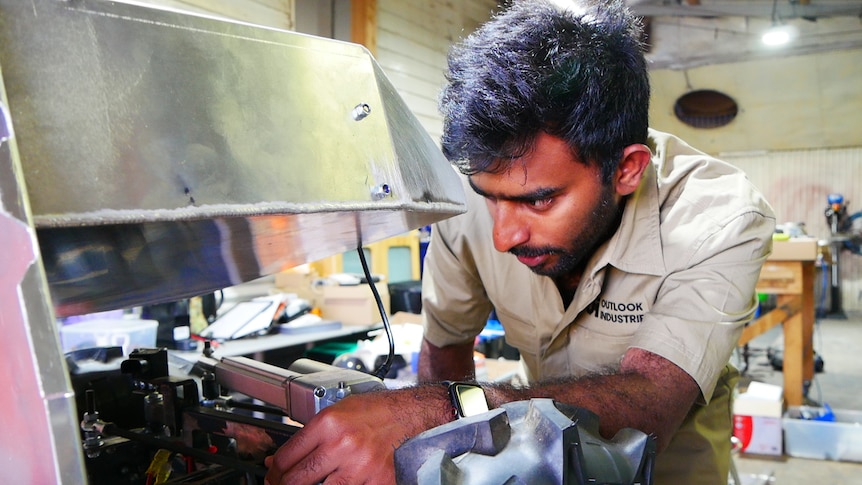 A bearded man in a khaki shirt leaning over some machinery to work on it