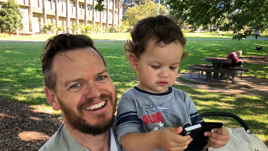 Smiling father Michael Fogarty holds his young song Ethan as he plays on a smart phone, in a park setting.