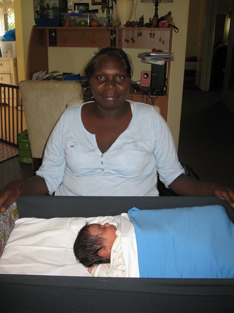 Aboriginal mum sits smiling behind her baby who is sleeping in a Pepi-Pod