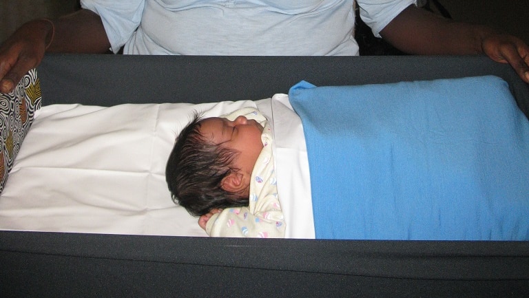 Aboriginal mum sits smiling behind her baby who is sleeping in a Pepi-Pod