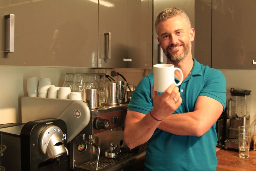 Man standing in kitchen holds up coffee mug.