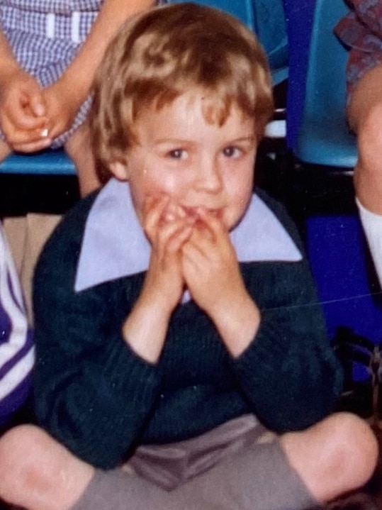 A close-up shot of a young schoolboy sitting with legs crossed on the floor with his mouth partially covered by his hands.