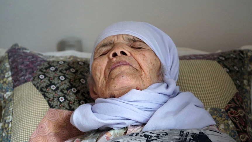 A close-up of an elderly woman lying in bed wearing a light blue head scarf.