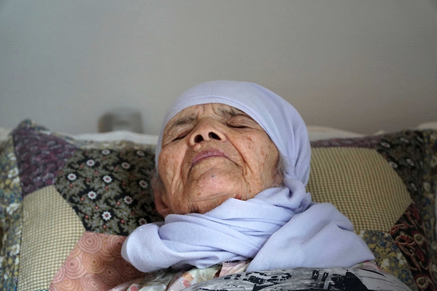 A close-up of an elderly woman lying in bed wearing a light blue head scarf.