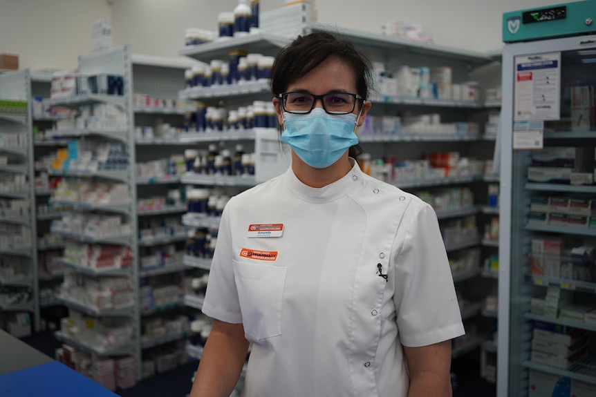A woman in a white labcoat wears a mask and glasses, standing in a pharmacy.