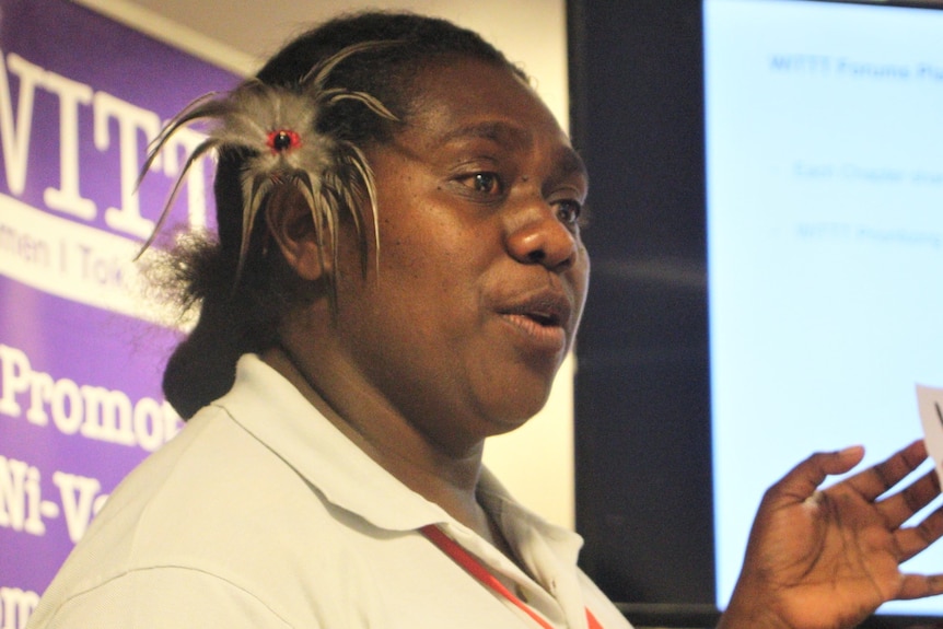 A ni-vanuatu woman giving a presentation with a slideshow in the background and a feather above her ear
