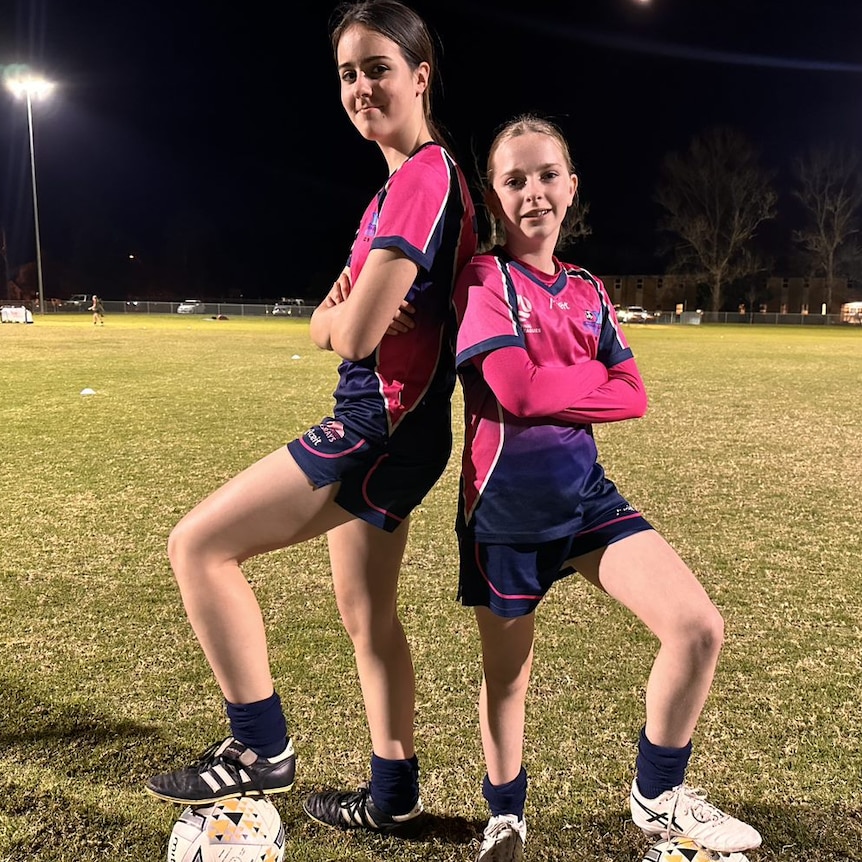 Two girls wearing soccer uniforms stand back to back with arms crossed.