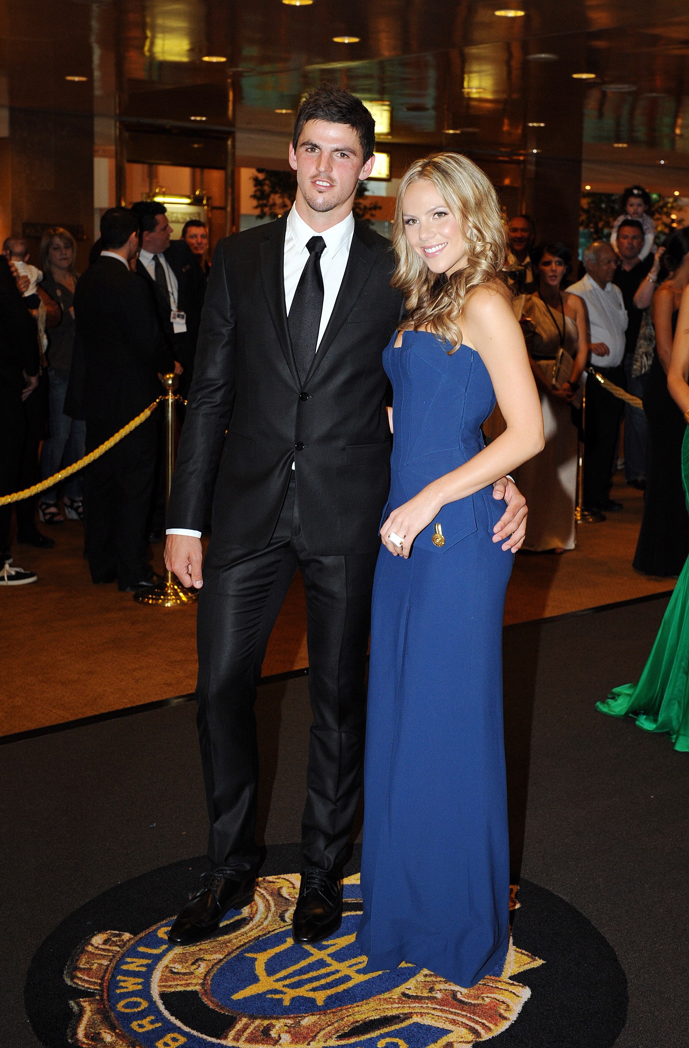 A man in a suit and a blonde woman in a blue sleeveless dress at a late-night event.